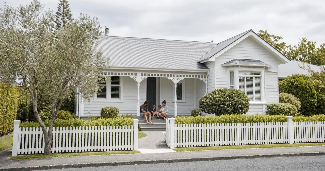 A family sitting together on the front steps of a white weatherboard villa, seen from the street.