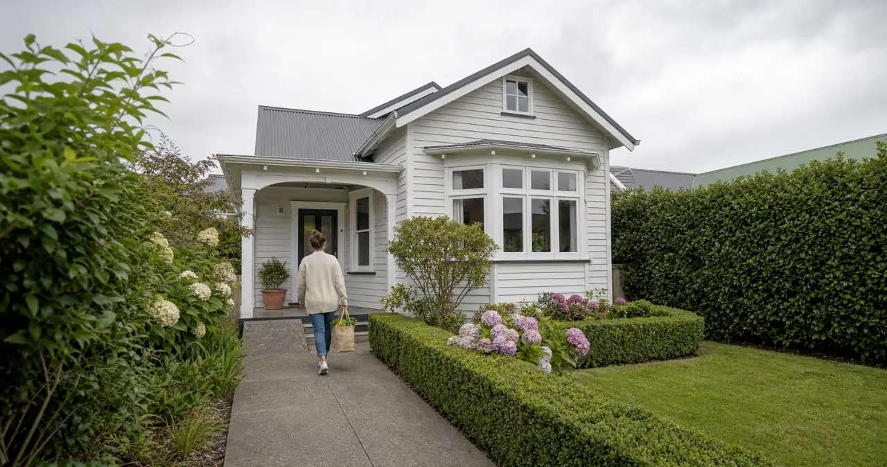 A person walking up the front path of a weatherboard bungalow, past hydrangeas and a clipped hedge.