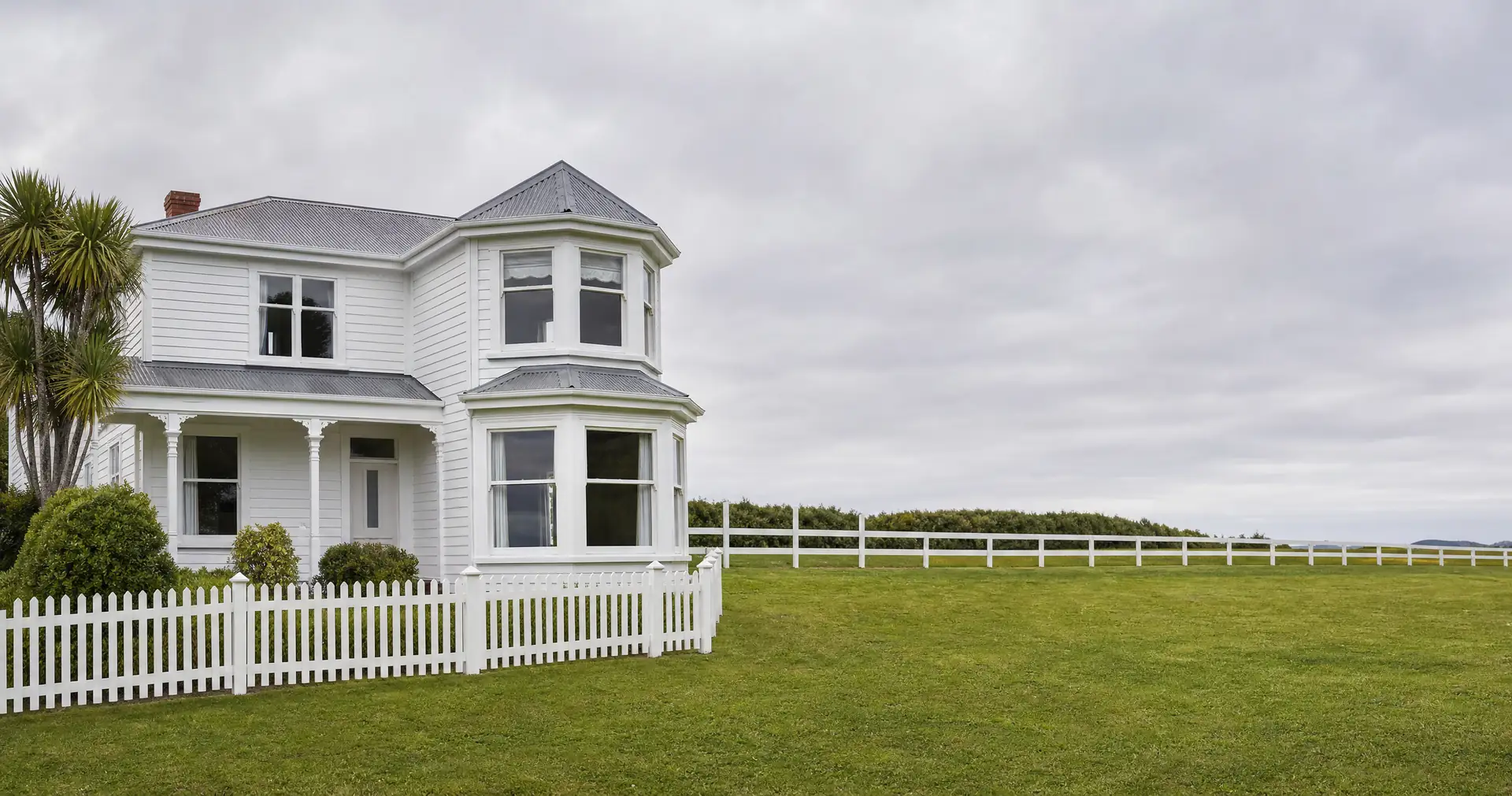 A two-storey white weatherboard villa with a bay window and a veranda. A white picket fence and a mown lawn sit in front.