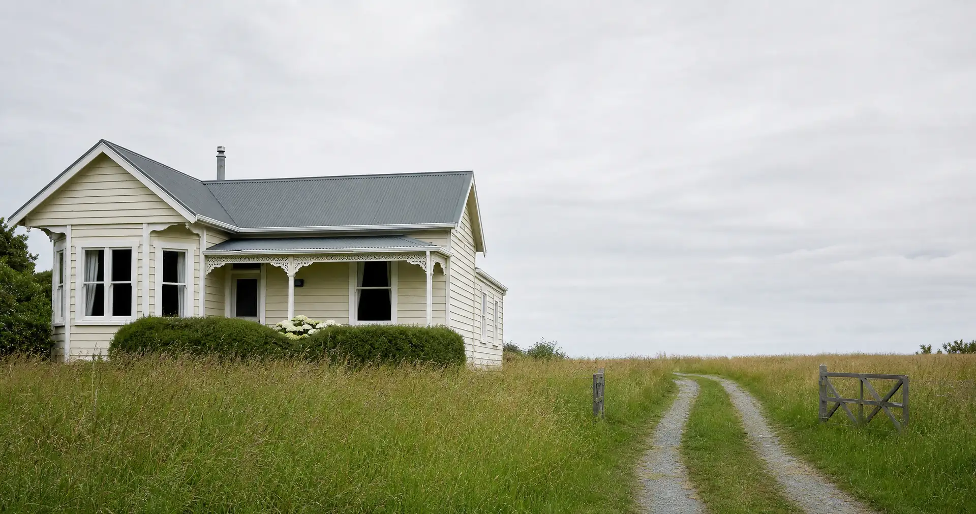 An empty cream weatherboard cottage with dark, blank windows. The grass is long and the gate stands open.