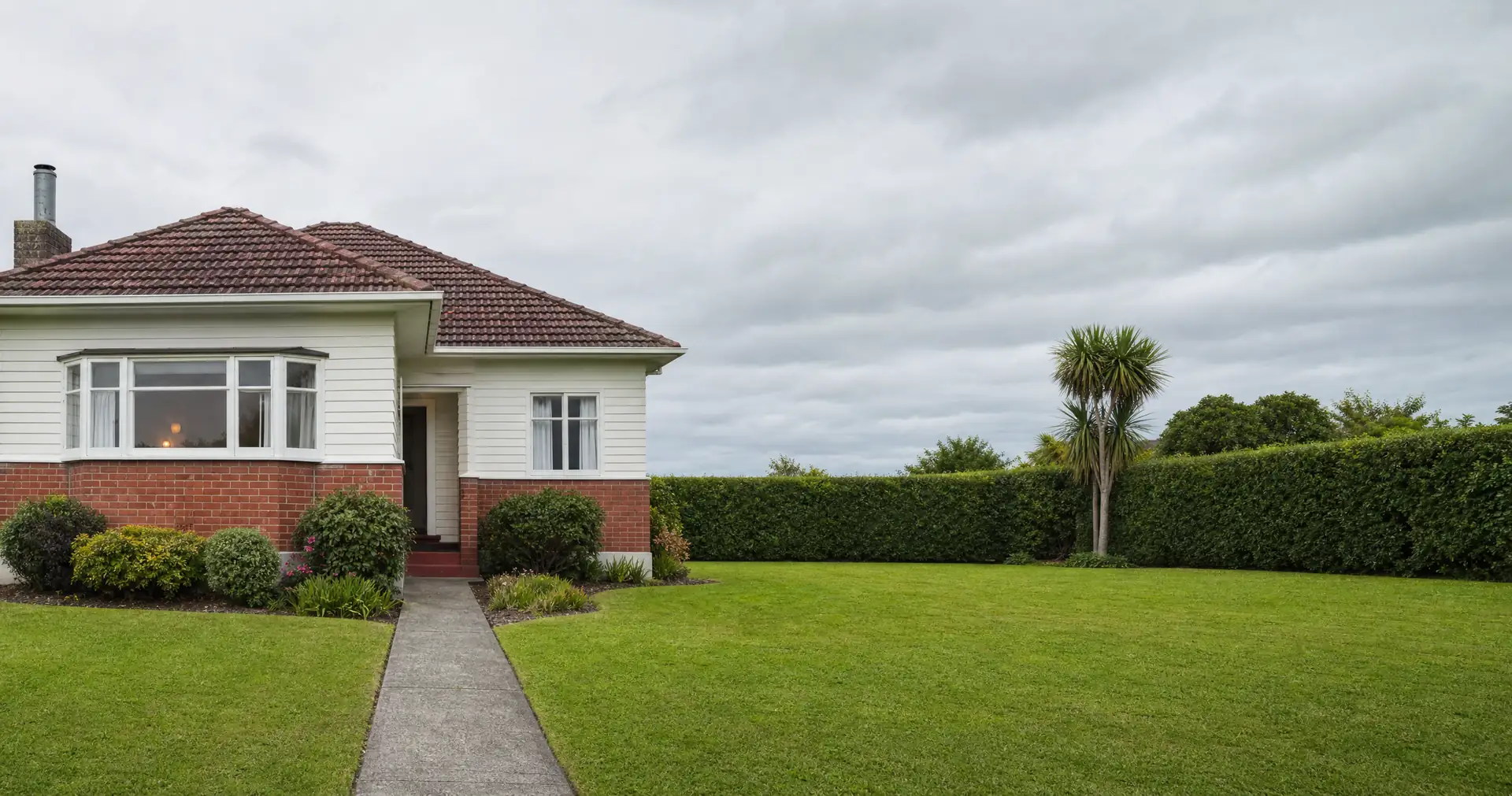 A nineteen-forties state house, brick at the bottom and weatherboard above. A concrete path crosses a wide front lawn.
