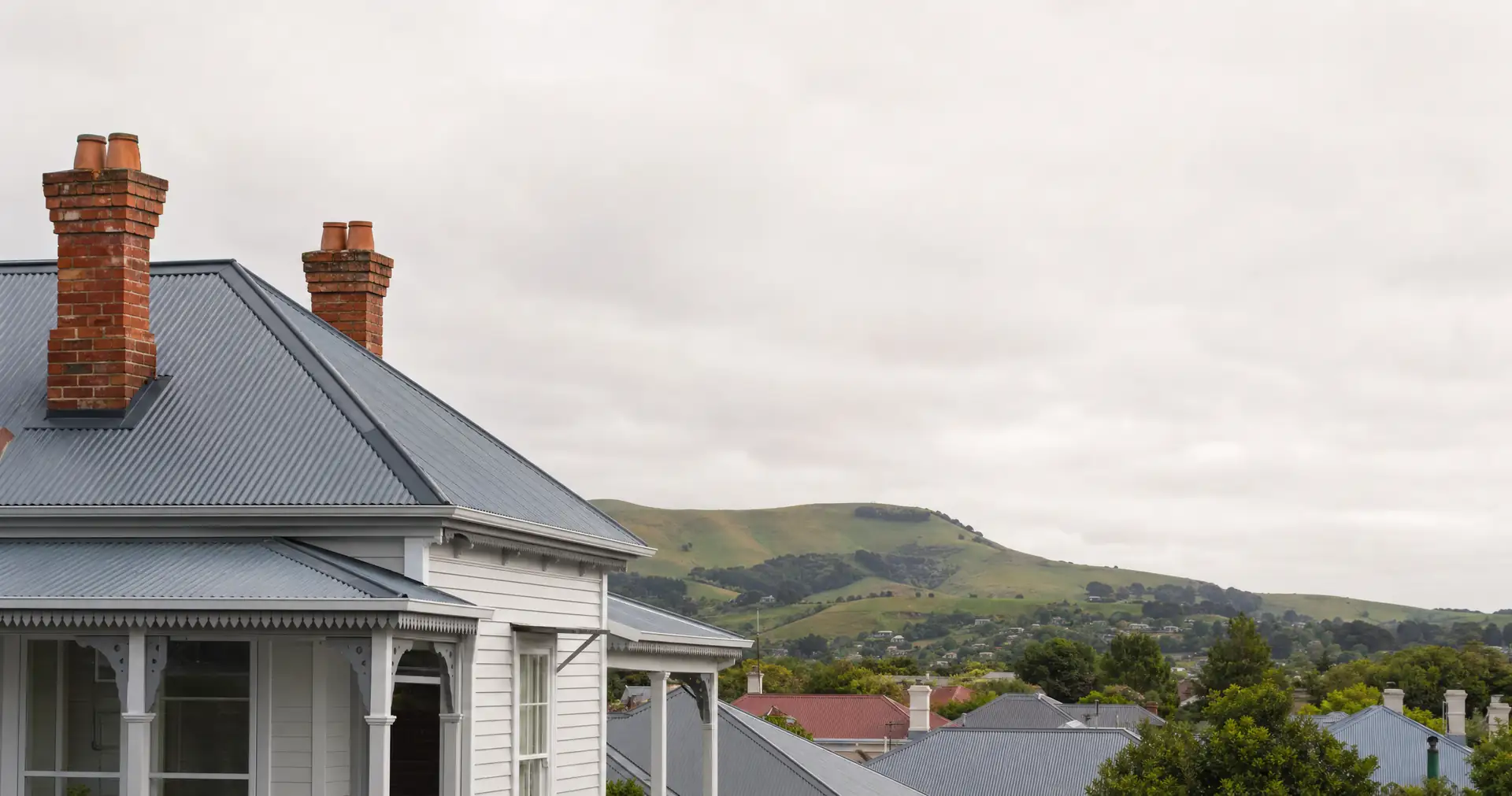 Corrugated iron rooflines and brick chimneys on older weatherboard houses, with green hills behind.