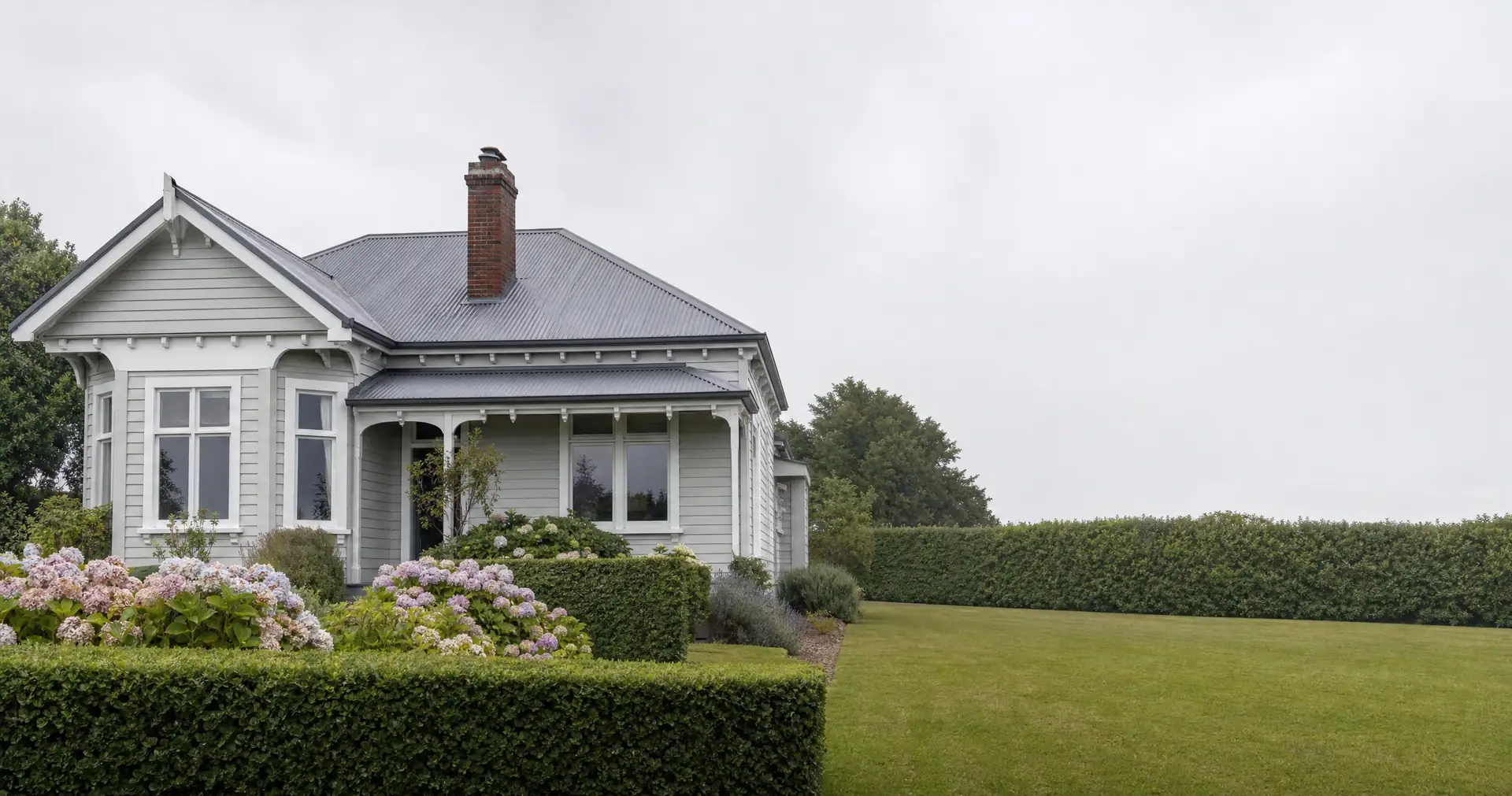 A grey weatherboard bungalow with a corrugated iron roof and a brick chimney. A clipped hedge and hydrangeas line the path.