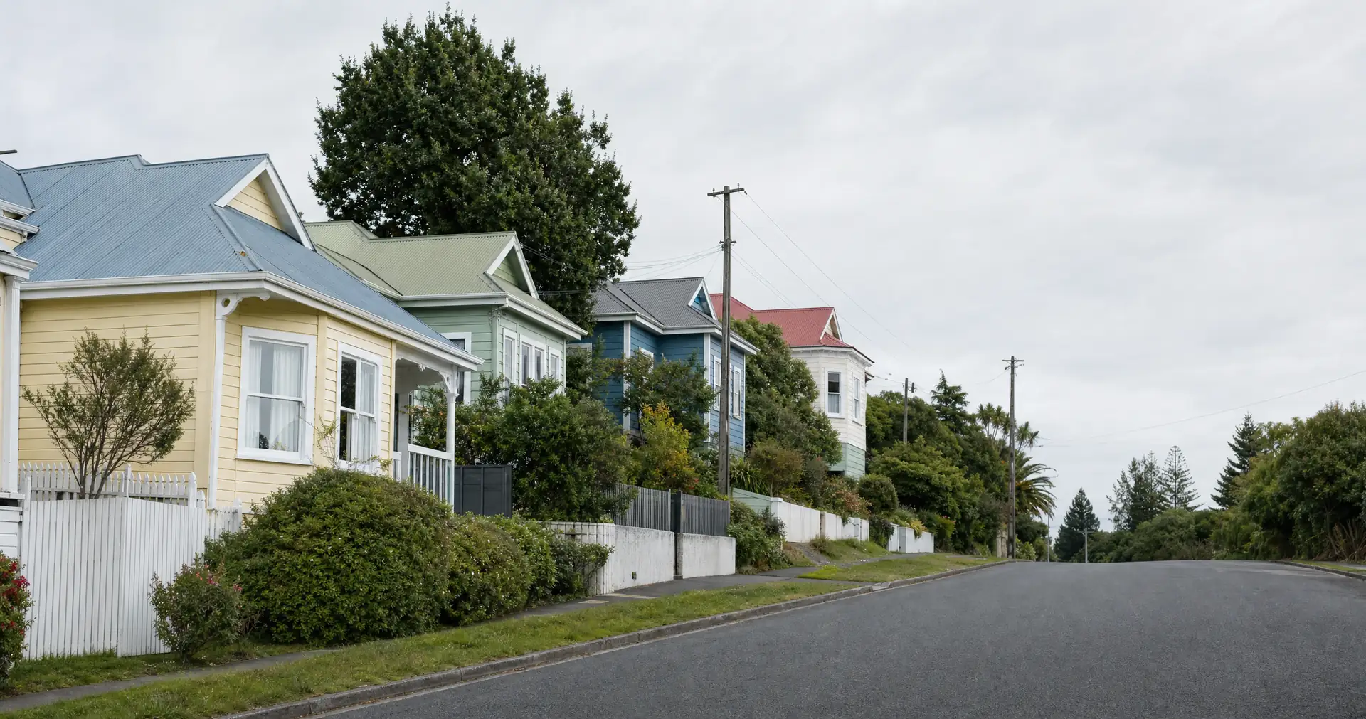 A row of weatherboard houses in yellow, green and blue. They step up a suburban hill behind hedges and power poles.